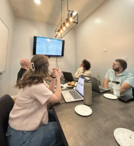 Storey Marketing team members gather around a meeting table during an April team meeting, reviewing information displayed on a television screen while discussing marketing, web development, and pharmacy industry initiatives.